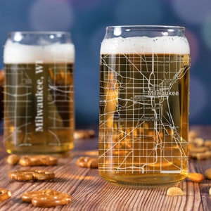 May include: Two clear glass beer mugs with a white outline of the city of Milwaukee, Wisconsin. The mug on the right is filled with a light amber beer. The mugs are sitting on a wooden table with pretzels and nuts.