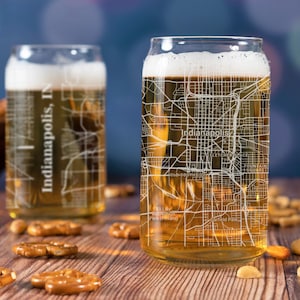 May include: Two clear glass beer can glasses with a white outline of the city of Indianapolis, Indiana. The glasses are filled with a light amber colored beer. The glass on the right has a white outline of the city streets and landmarks. The glass on the left has the text "Indianapolis, IN" on the side.