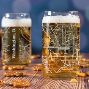 May include: Two clear glass beer mugs with a white outline of the Washington, D.C. map etched on the side. The mugs are filled with a light amber colored beer. The mugs are sitting on a wooden table with pretzels scattered around them.
