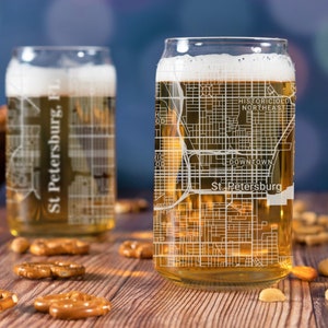 May include: Two clear glass beer mugs with a map of St. Petersburg, Florida etched on the side. The mugs are filled with a golden-colored beverage and are sitting on a wooden table with pretzels and nuts.