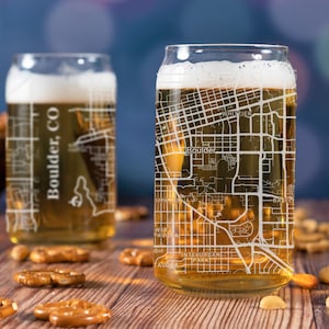 May include: Two clear glass beer mugs with a map of Boulder, CO etched on the side. The mugs are filled with beer and sitting on a wooden table with pretzels and nuts.
