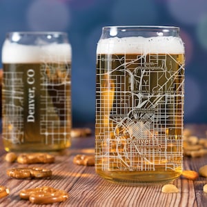 May include: Two clear glass beer mugs with a white outline of the Denver, Colorado street map. The mugs are filled with a golden-colored liquid and are sitting on a wooden table with pretzels and nuts.