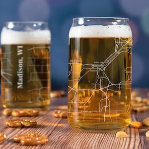 May include: Two clear glass beer mugs with a map of Madison, WI etched on the side. The mug on the right is filled with a light golden beer. The mugs are sitting on a wooden surface with pretzels and nuts.