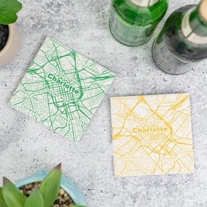May include: Two square coasters with a map of Charlotte, North Carolina. One coaster has a green map on a white background, and the other coaster has a yellow map on a white background.