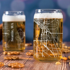 May include: Two clear glass beer mugs with a white outline of the Seattle, Washington city map printed on the side. The mugs are filled with a light amber colored beer. The mugs are sitting on a wooden table with pretzels and nuts scattered around them.