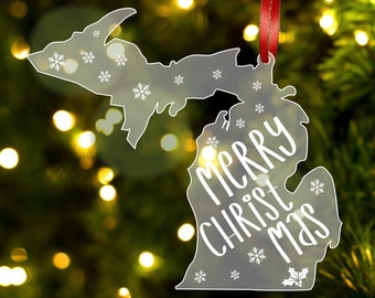 May include: A clear acrylic Christmas ornament shaped like the state of Michigan with the words "Merry Christmas" written inside. The ornament has a red ribbon attached to the top.
