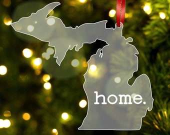 May include: A clear acrylic Christmas ornament shaped like the state of Michigan with the word "home" printed in white on the lower right side. The ornament is hanging from a red ribbon.