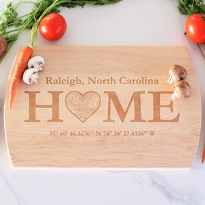 May include: A wooden cutting board with the words "Raleigh, North Carolina" and "HOME" engraved on it. The word "HOME" has a heart shape in the middle with an outline of the city of Raleigh. The coordinates "35° 46' 46.5276" N 78° 38' 17.4336" W" are also engraved on the board.