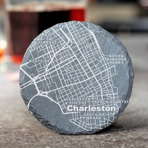 May include: A gray slate coaster with a white map of Charleston, South Carolina. The map includes the names of neighborhoods like Ansonborough Homes, Harleston Village, and French Quarter.