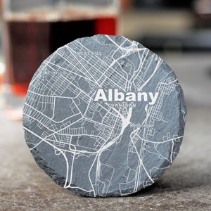 May include: A gray slate coaster with a white outline map of Albany, New York. The map includes the words "Albany", "Downtown", "Pine Hills", "West Hill", and "Rensselaer".