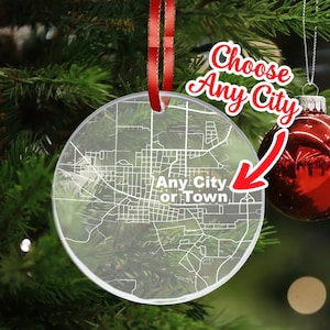 May include: A clear acrylic Christmas ornament with a white outline of a city map. The ornament is hanging from a red ribbon and has the text "Choose Any City or Town" printed on it.