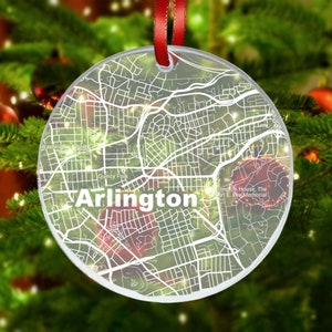 May include: A clear, round Christmas ornament with a map of Arlington, Virginia, in white. The word "Arlington" is prominently displayed. A red ribbon is attached for hanging. The ornament is set against a backdrop of a Christmas tree.