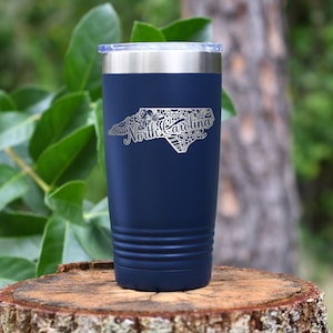 May include: A navy blue insulated tumbler with a silver lid. The tumbler features a detailed silver outline of the North Carolina state map, with the state name in elegant script. The tumbler is cylindrical with a ridged design near the base.