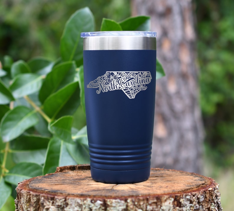 May include: A navy blue insulated tumbler with a silver lid. The tumbler features a detailed silver outline of the North Carolina state map, with the state name in elegant script. The tumbler is cylindrical with a ridged design near the base.