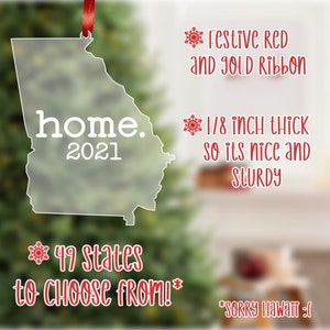 May include: A clear acrylic ornament shaped like the state of Georgia with the word "home" and the year "2021" printed on it. The ornament is decorated with a red ribbon and a snowflake. The text "49 states to choose from!" is printed below the ornament.