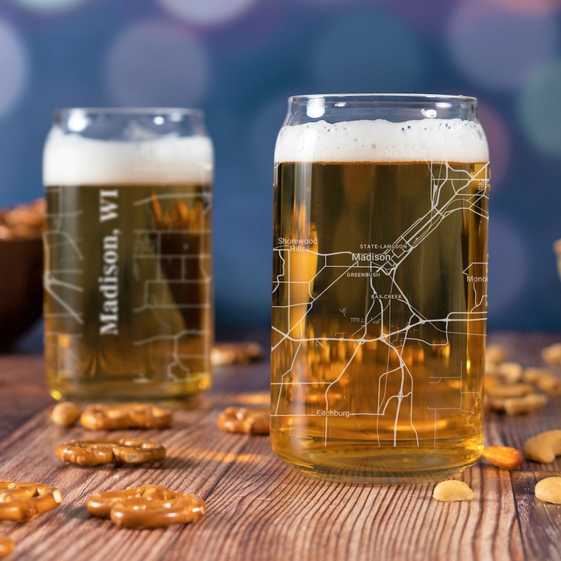 May include: Two clear glass beer mugs with a map of Madison, WI etched on the side. The mug on the right is filled with a light golden beer. The mugs are sitting on a wooden surface with pretzels and nuts.