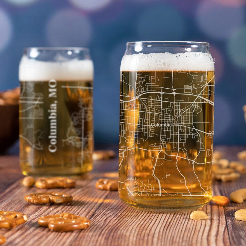 May include: Two clear glass beer can glasses with a map of Columbia, MO etched on the side. The glasses are filled with a golden beer and are sitting on a wooden table with pretzels and nuts.