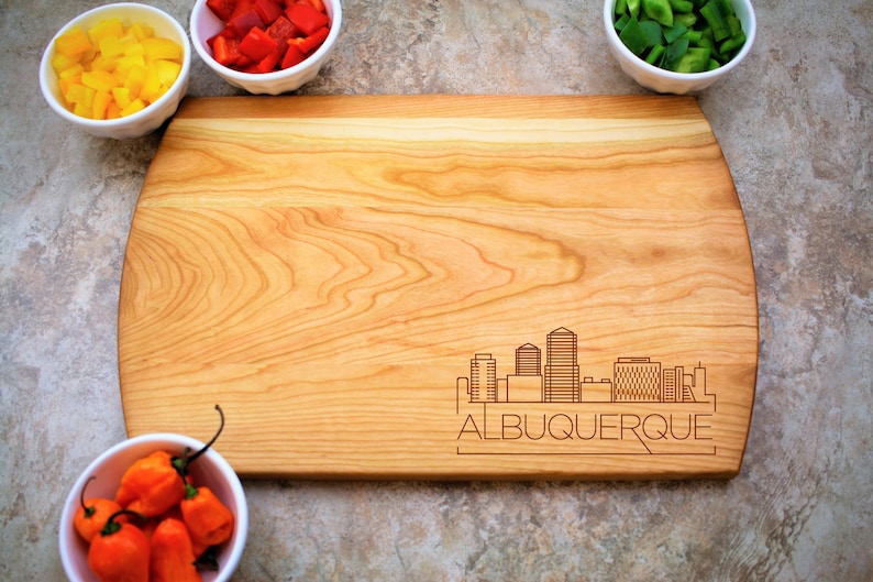 Pode incluir: Uma t&aacute;bua de cortar de madeira com um design de cidade e o texto "ALBUQUERQUE" gravado na parte inferior. A t&aacute;bua &eacute; cercada por tigelas de vegetais picados.