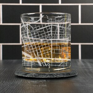 May include: A clear glass tumbler with a map of Tuscaloosa, Alabama etched on the side. The glass is filled with amber liquid and is sitting on a black slate coaster.