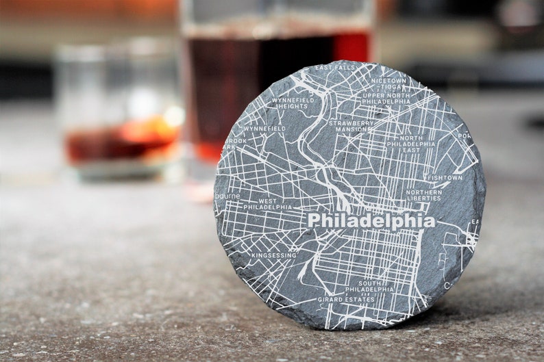 May include: A round, gray stone coaster with a white map of Philadelphia, Pennsylvania. The map includes neighborhood names like Fishtown, Northern Liberties, and South Philadelphia.