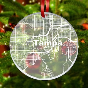 May include: A clear glass Christmas ornament with a white outline map of Tampa, Florida. The ornament features the text "Tampa" and various neighborhood names.