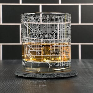 May include: A clear glass rocks glass with an etched map of Washington, D.C. The glass is filled with amber-colored liquid and is sitting on a black slate coaster.