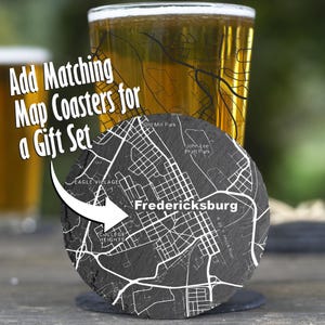 May include: A round, dark gray slate coaster featuring a white engraved street map of Fredericksburg, including labels for Eagle Village and College Heights. The text Add Matching Map Coasters for a Gift Set is overlaid on the image.