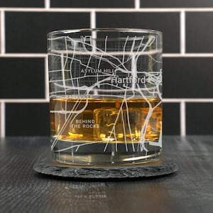 May include: A clear glass with a map of Hartford, Connecticut etched on the side. The glass is filled with amber liquid and ice cubes. The glass is sitting on a black slate coaster.