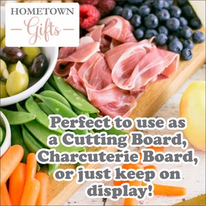 Pode incluir: Uma t&aacute;bua de cortar de madeira com uma variedade de alimentos, incluindo prosciutto, azeitonas, feij&atilde;o verde, cenouras, mirtilos e framboesas. A t&aacute;bua &eacute; perfeita para servir charcutaria ou como pe&ccedil;a decorativa. O texto "HOMETOWN -Gifts- Perfect to use as a Cutting Board, Charcuterie Board, or just keep on display!" est&aacute; impresso na t&aacute;bua.