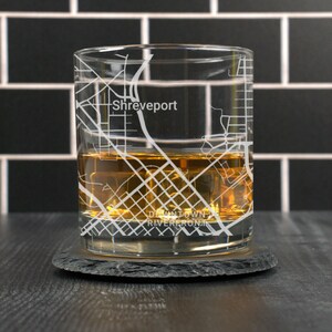 May include: A clear glass with an etched design of the Shreveport, Louisiana city map. The glass is filled with amber-colored liquid and ice cubes. The text "Downtown Shreveport" and "Riverfront" is visible at the bottom of the glass.