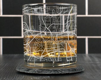 May include: A clear glass with a design of the Winston-Salem city map etched into the side. The glass is filled with amber liquid and is sitting on a black slate coaster.