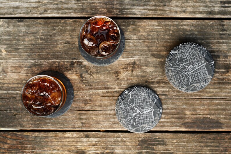 May include: Three black slate coasters with a white outline of the city of Philadelphia. Two coasters have a glass of iced tea on top of them.