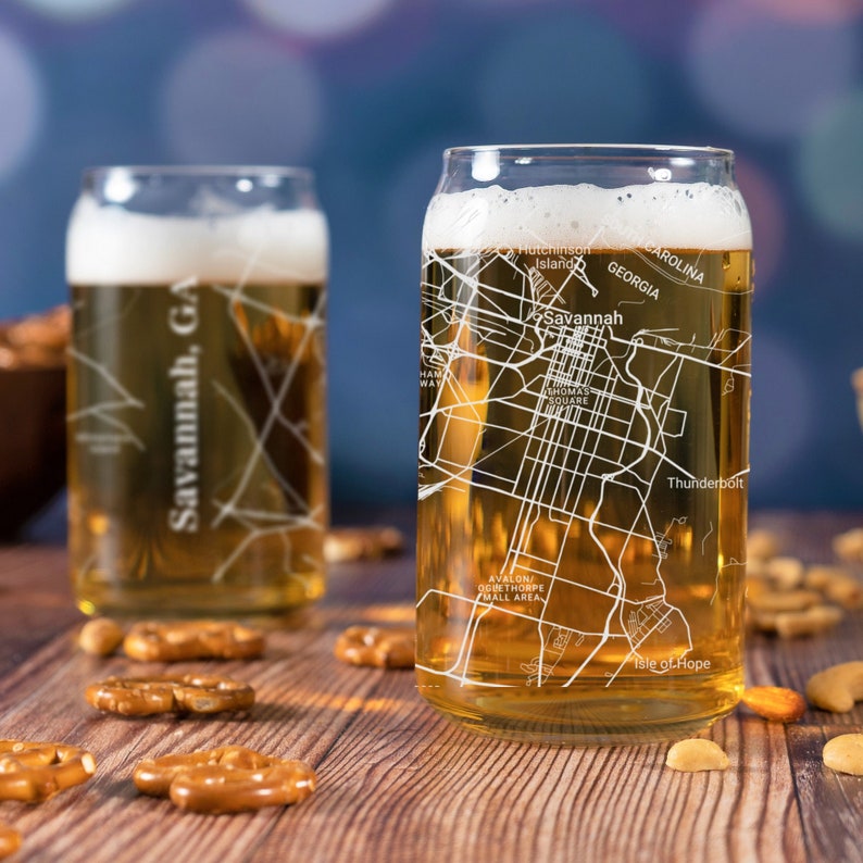 May include: Two clear glass beer can glasses with a white outline of the city of Savannah, Georgia printed on them. The glasses are filled with a light amber colored beer. The glasses are sitting on a wooden table with pretzels scattered around them.