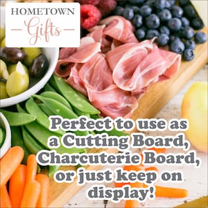 May include: A wooden cutting board with a variety of food items arranged on it, including prosciutto, olives, green beans, carrots, blueberries, and raspberries. The board is labeled "HOMETOWN -Gifts- Perfect to use as a Cutting Board, Charcuterie Board, or just keep on display!"