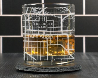 May include: A clear glass rocks glass with a map of Ann Arbor, Michigan etched on the side. The glass is filled with amber liquid and ice cubes. The glass is sitting on a black slate coaster.