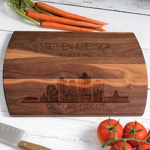 May include: A personalized walnut wood cutting board with a laser engraved cityscape of Salt Lake City, Utah. The board is engraved with the names "Stephen & Jessica" and the date "October 18, 2022".