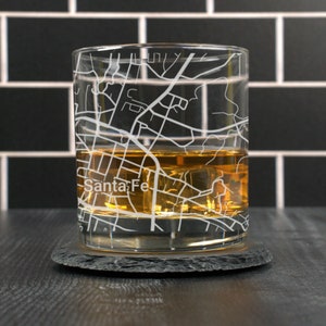 May include: A clear glass rocks glass with a white outline of the city of Santa Fe, New Mexico, etched into the side. The glass is filled with amber-colored liquid and ice cubes. The glass is sitting on a black slate coaster.