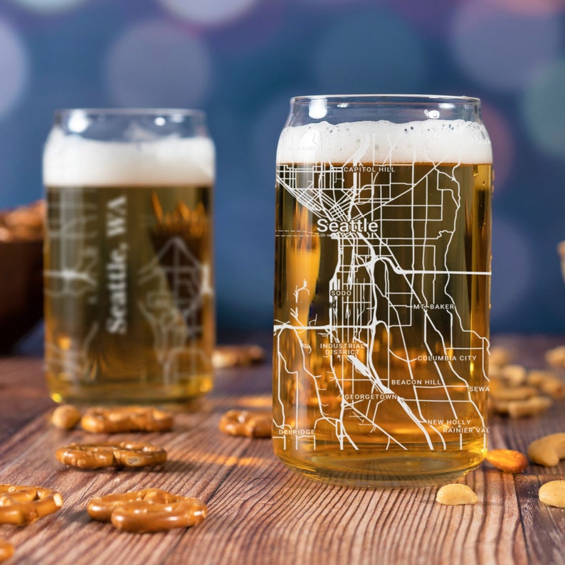 May include: Two clear glass beer mugs with a white outline of the Seattle, Washington city map printed on the side. The mugs are filled with a light amber colored beer. The mugs are sitting on a wooden table with pretzels and nuts scattered around them.