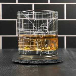 May include: A clear glass with a design of the Milwaukee city map etched on the side. The glass is filled with amber liquid and ice cubes. The glass is sitting on a black slate coaster.