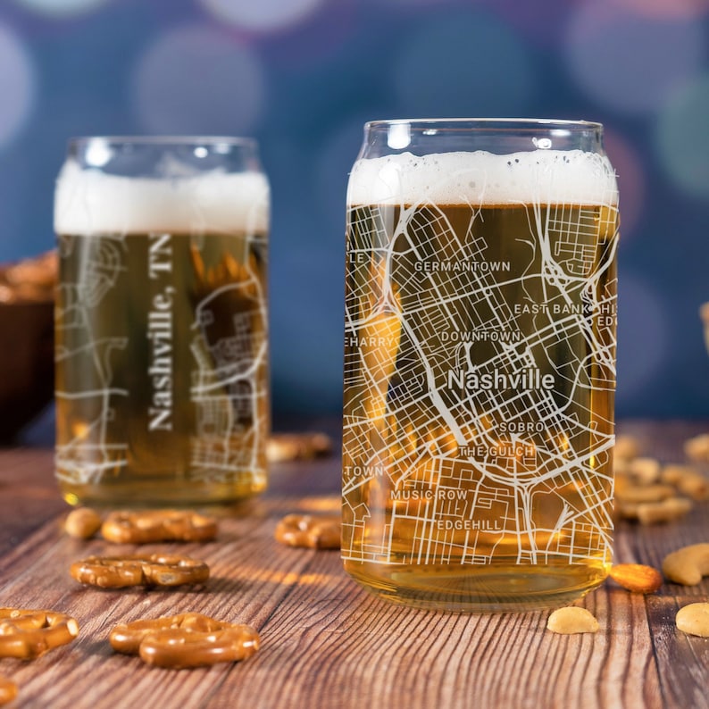 May include: Two clear glass beer mugs with a white outline of the city of Nashville, Tennessee. The mugs are filled with a golden-colored beverage and are sitting on a wooden surface with pretzels.
