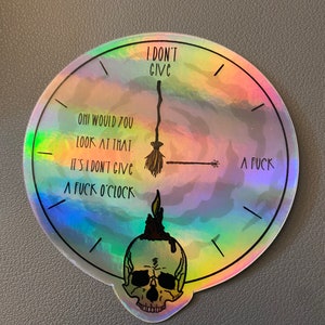 Op de afbeelding: Holografische sticker in de vorm van een klok met de tekst "I DON'T GIVE A FUCK O'CLOCK". De wijzers van de klok zijn een bezem en een toverstaf. Onderaan staat een schedel met een kaars.