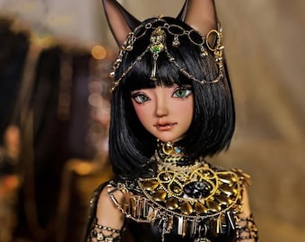 May include: A black and gold doll with cat ears and a black wig. The doll is wearing a black and gold costume with a large gold necklace. The doll has green eyes and a neutral expression.