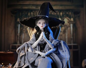 May include: A porcelain doll wearing a black and grey dress and a black witch's hat. The doll is sitting on a chair with a dark background.