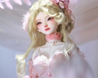 May include: A porcelain doll with blonde hair and delicate facial features. The doll wears a pink dress with lace trim and a matching choker. A pink flower and feathers adorn the doll's hair.
