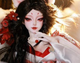 May include: A detailed doll with a fox-like appearance, featuring white ears, red eyes, and dark hair. The doll wears a red and white patterned outfit and a pearl necklace with a red bead. Red floral accents adorn the hair.