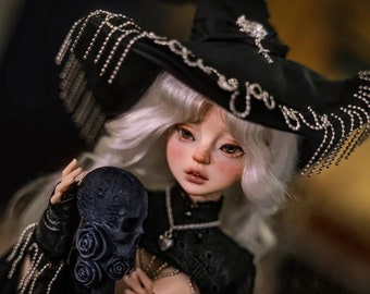 May include: A white-haired doll wearing a black witch's hat with silver embellishments and a black lace dress. The doll is holding a black skull with roses.