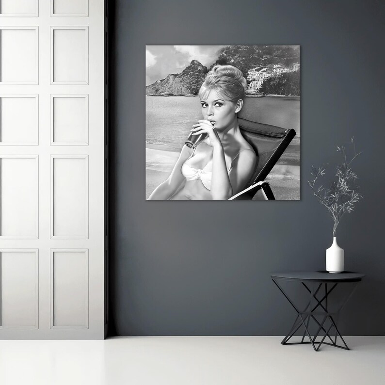 Puede incluir: Fotograf&iacute;a en blanco y negro de una mujer en bikini blanco, sentada en una silla de playa, bebiendo de un vaso. El fondo presenta una playa, monta&ntilde;as y un pueblo costero. La imagen es una impresi&oacute;n de estilo vintage.