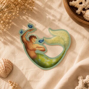 Puede incluir: Una pegatina troquelada que muestra a un tritón de pelo castaño con una cola verde moteada. El tritón aparece con los brazos levantados y rodeado de tres pequeños peces azules. La ilustración utiliza una paleta de colores suaves con tonos verde claro y amarillo.