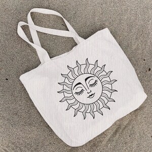 May include: A white canvas tote bag with a black line drawing of a sun with a face and closed eyes. The sun has a wavy, stylized design.