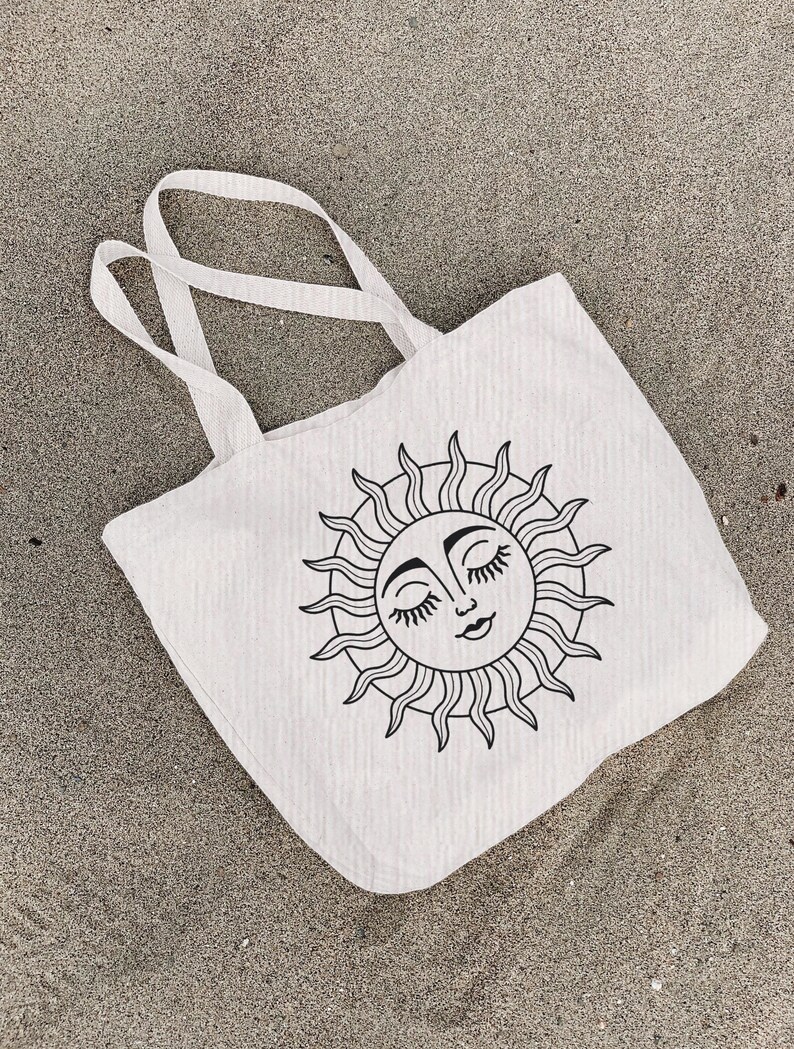 May include: A white canvas tote bag with a black line drawing of a sun with a face and closed eyes. The sun has a wavy, stylized design.